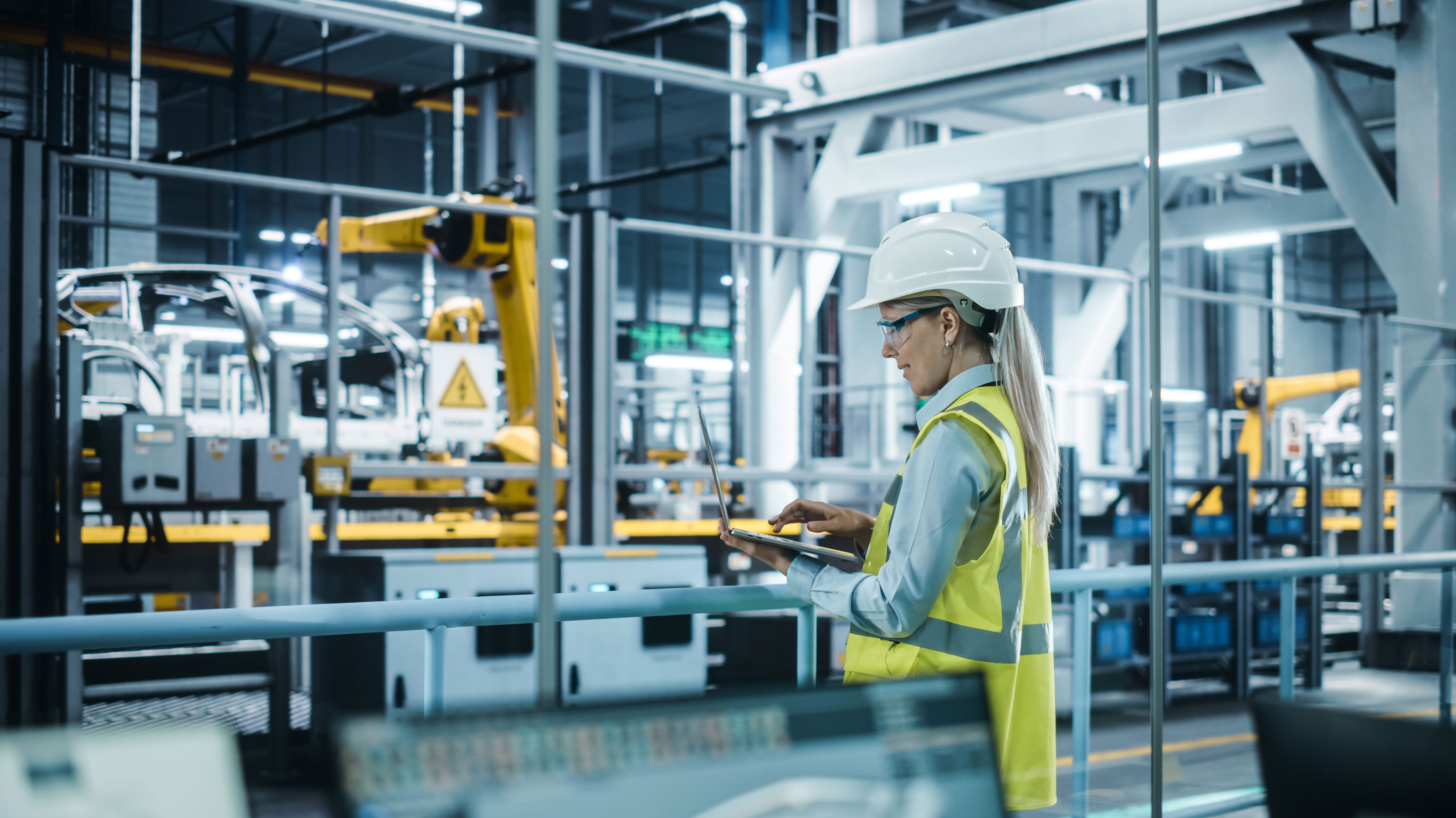 A female engineer wearing a hard hat and a safety vest works on a laptop while overlooking an automated factory floor with industrial robots.