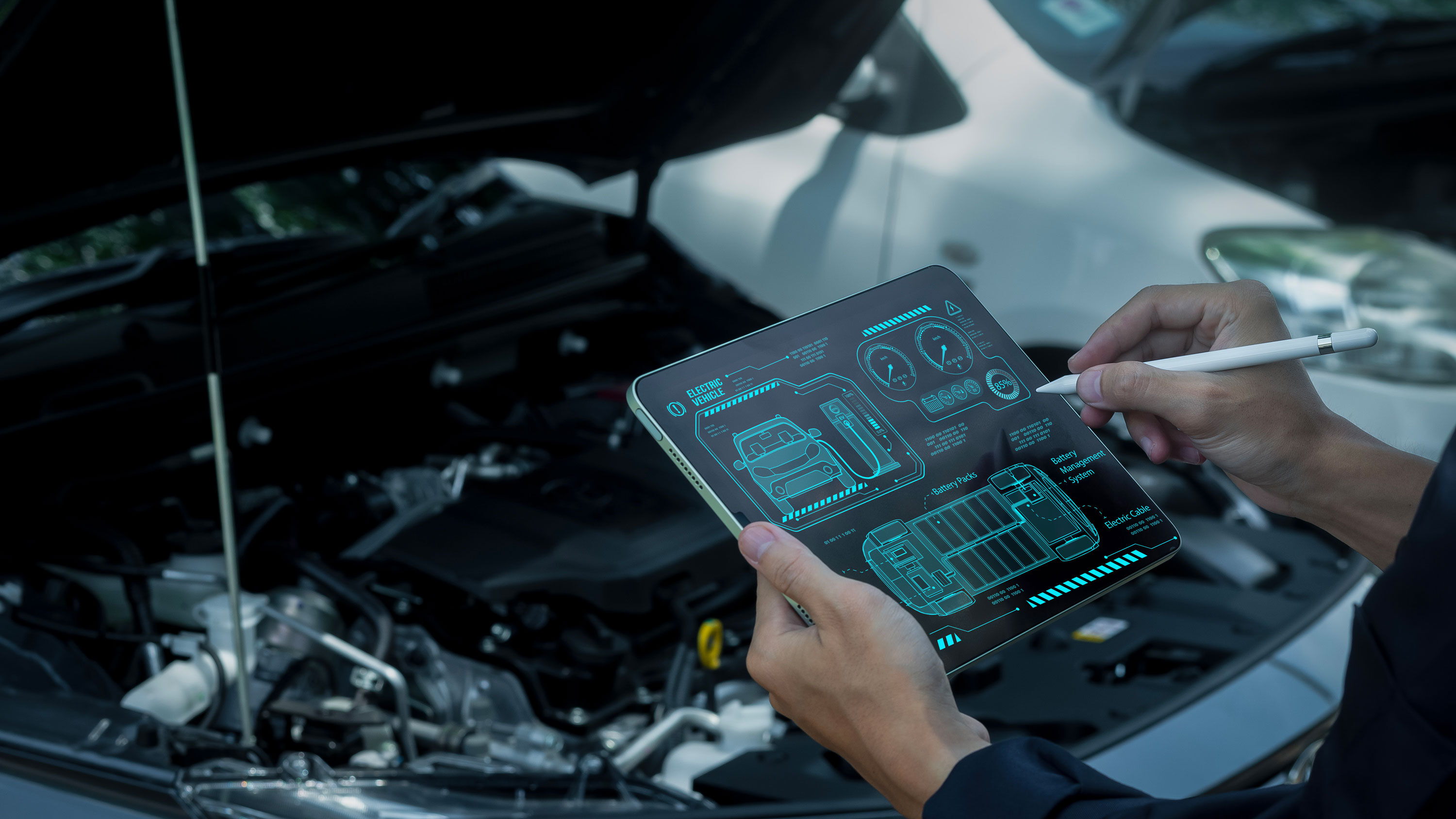 A technician holds a tablet displaying electric vehicle diagnostic software and uses a stylus while standing in front of a car with its hood open.