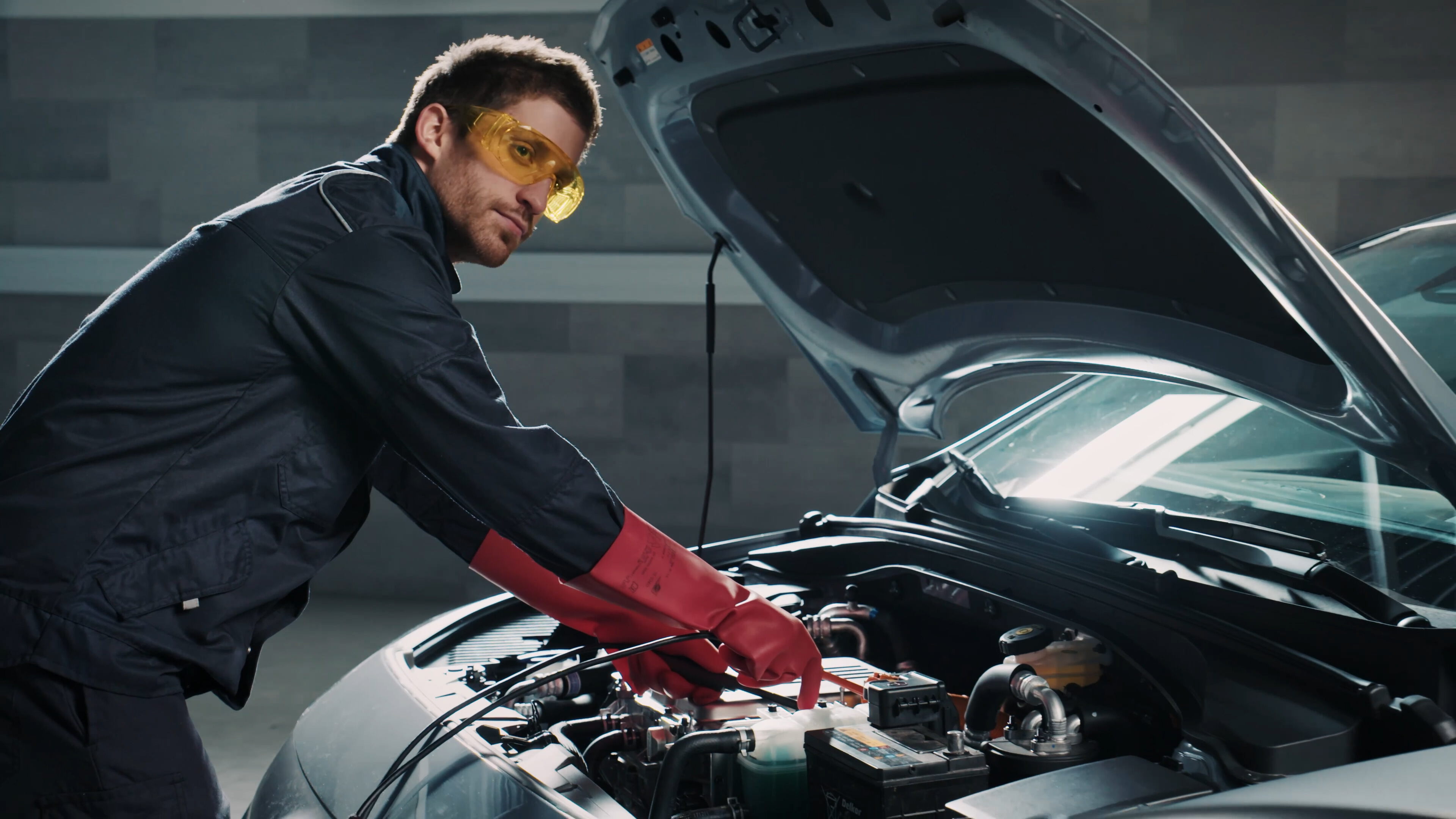 A car mechanic wearing yellow safety goggles and red gloves leans over the open hood of a car to work on the engine in a workshop.