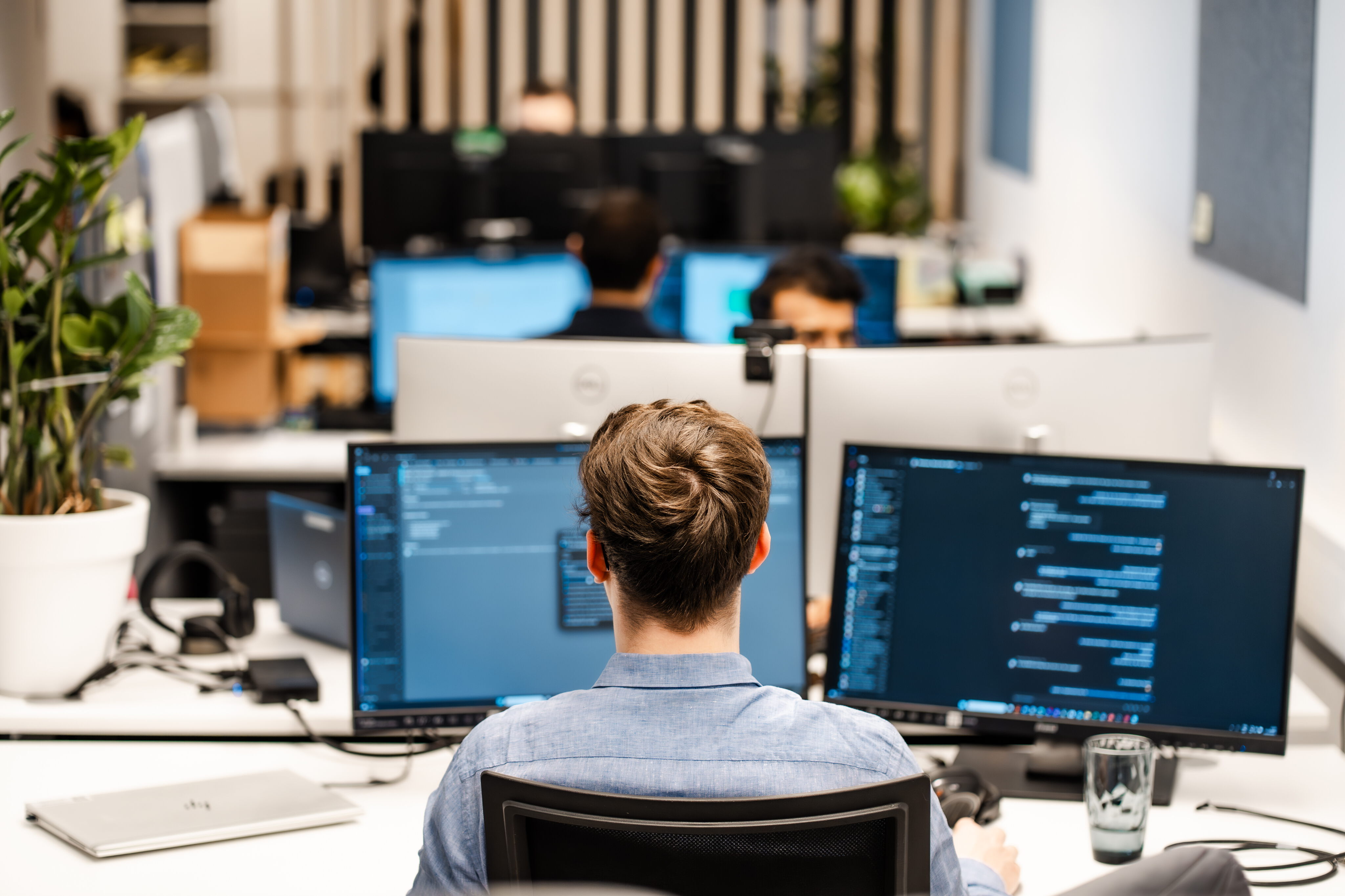 Rear view of a software developer sitting at a desk in an office, working on a dual-monitor computer setup displaying lines of code.