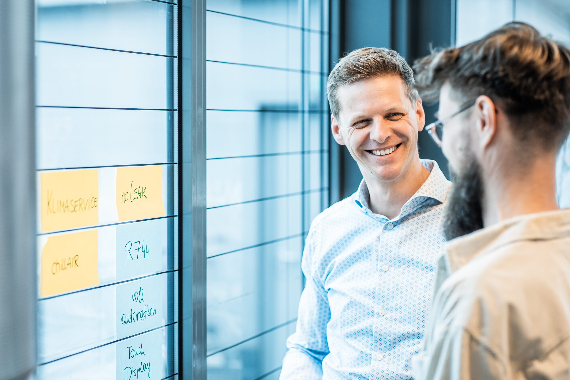A smiling man in a light blue shirt talks to his colleague in front of a glass wall covered with sticky notes.