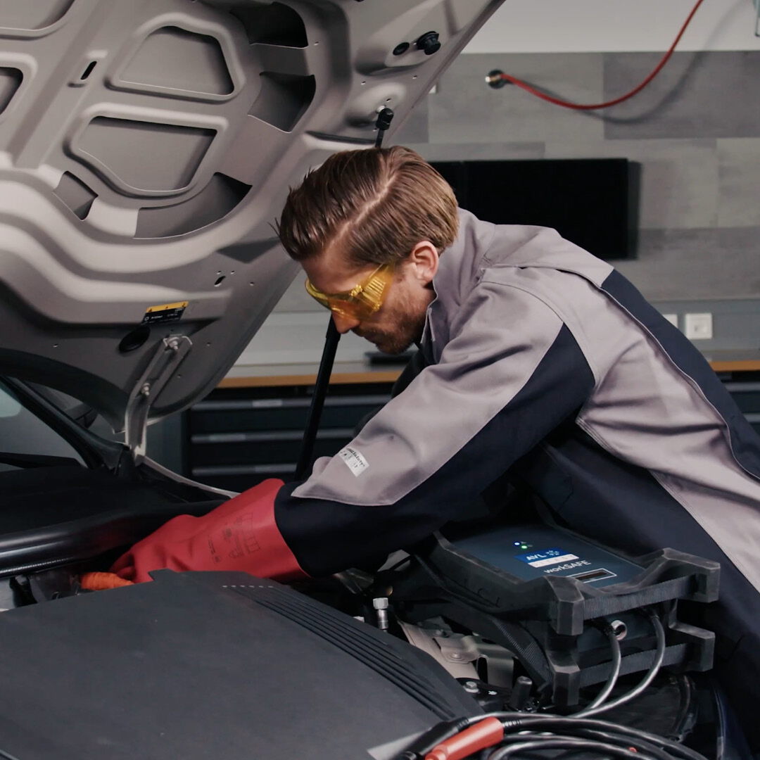Video thumbnail on the topic of safety: A female mechanic leans over the open engine bay of a car to work on it.