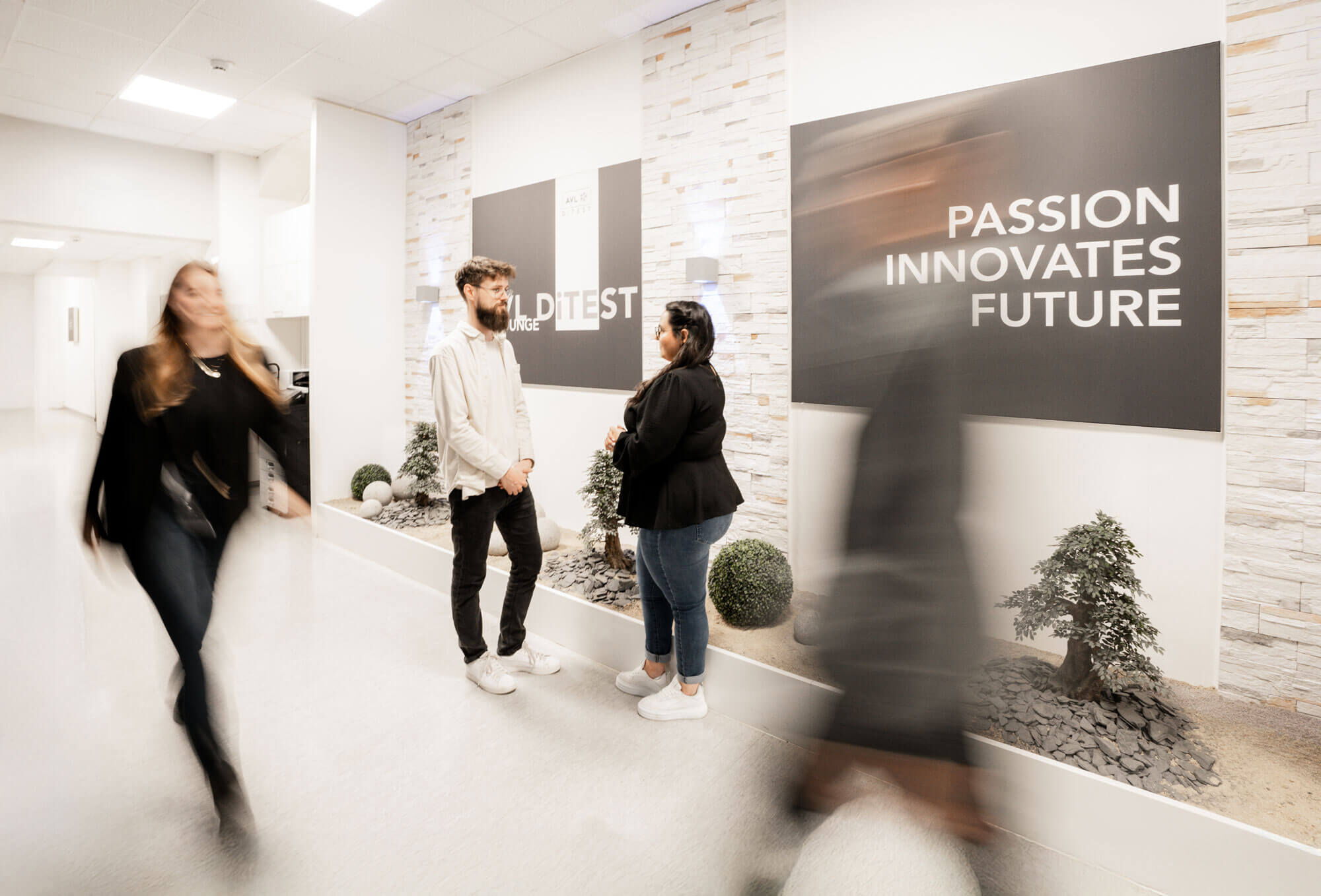 In a bright, modern office hallway, a man and a woman are standing and talking. Two other people walk past them with motion blur, creating a dynamic atmosphere. On the wall behind them is a large sign with the slogan "PASSION INNOVATES FUTURE".