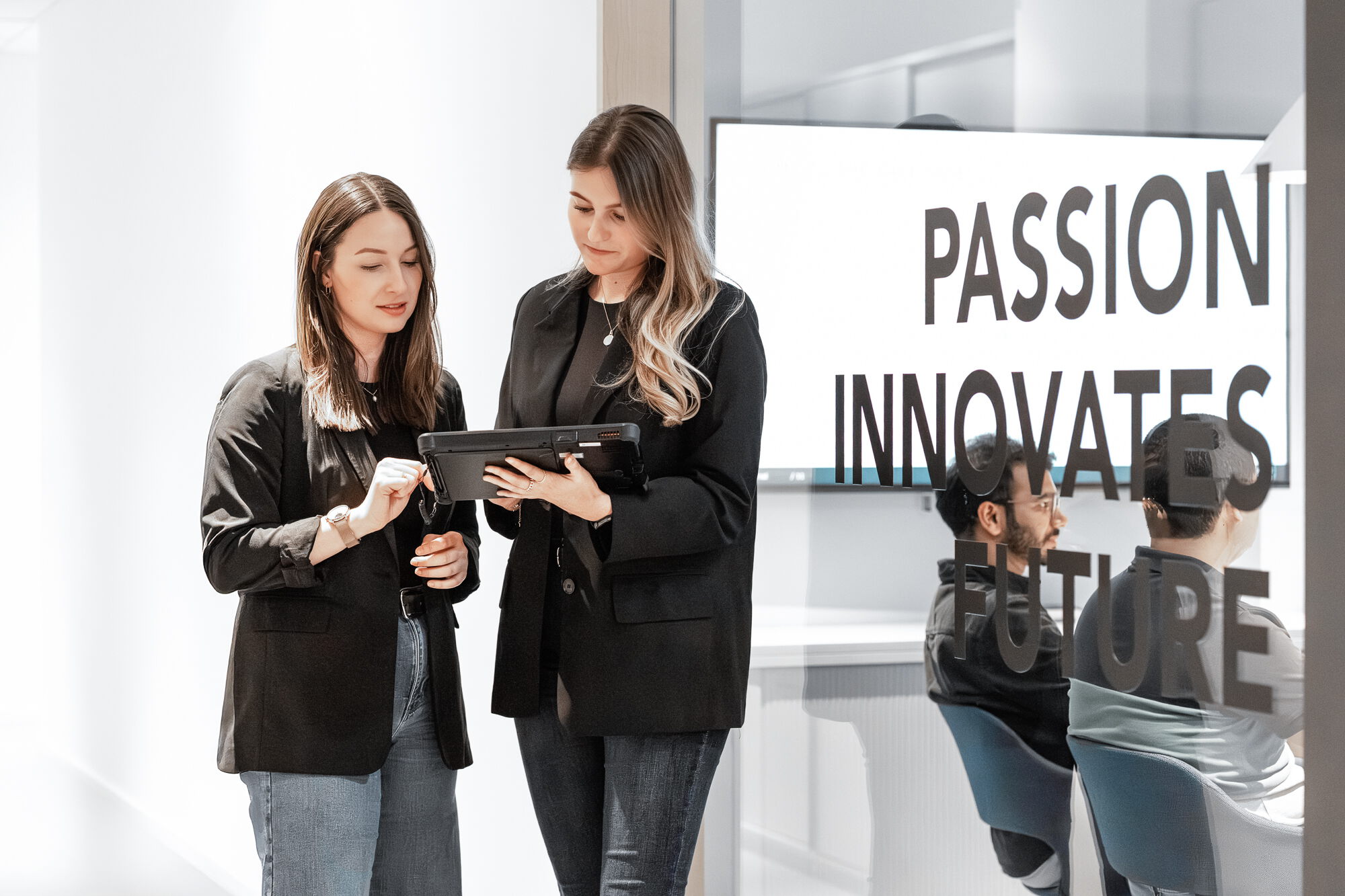 Two female colleagues in black blazers stand in a bright office hallway, smiling as they look at a tablet held by one of the women.