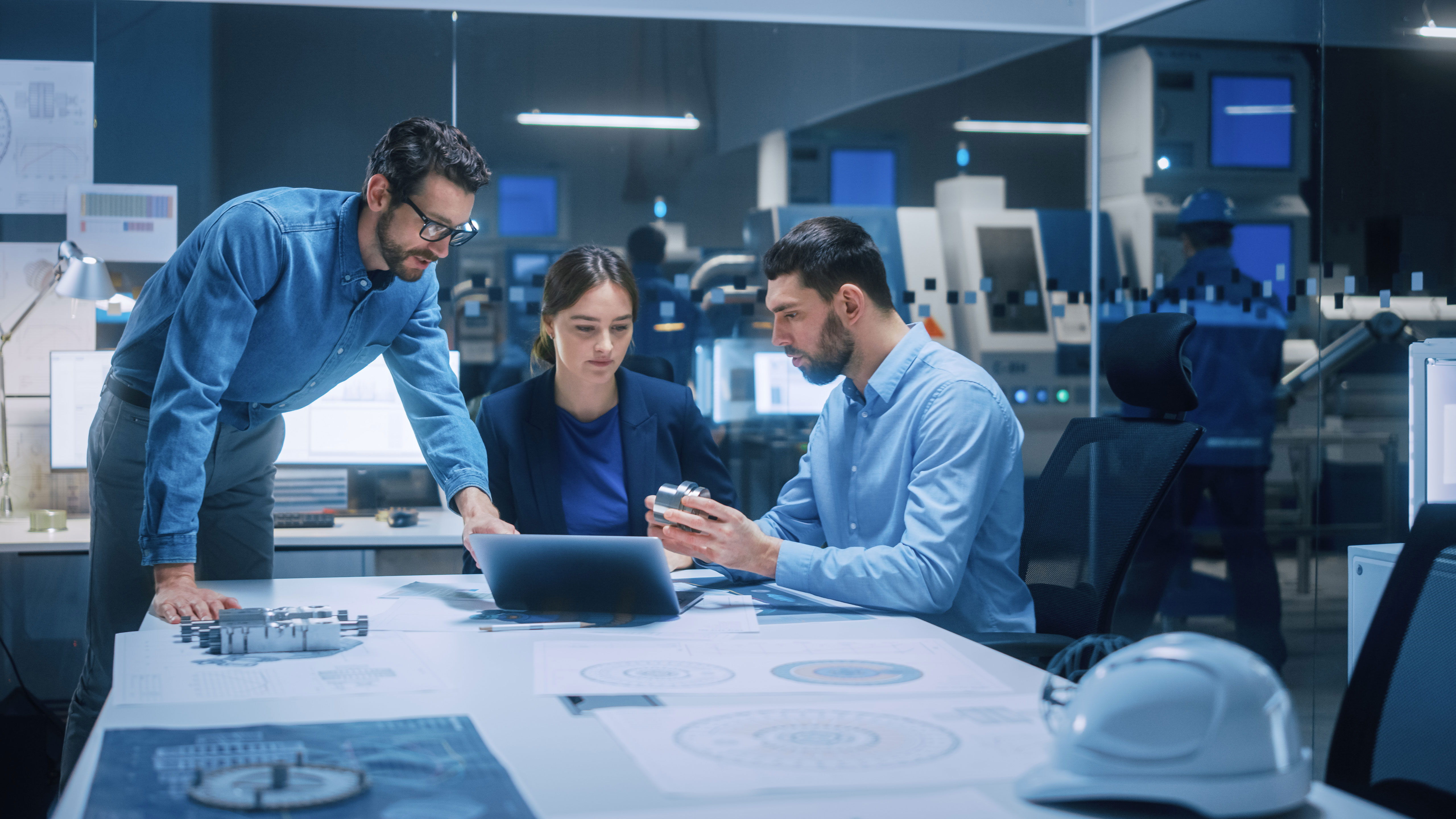 Three engineers gathered around a table with blueprints and a laptop, analyzing a metal prototype in a modern factory setting.